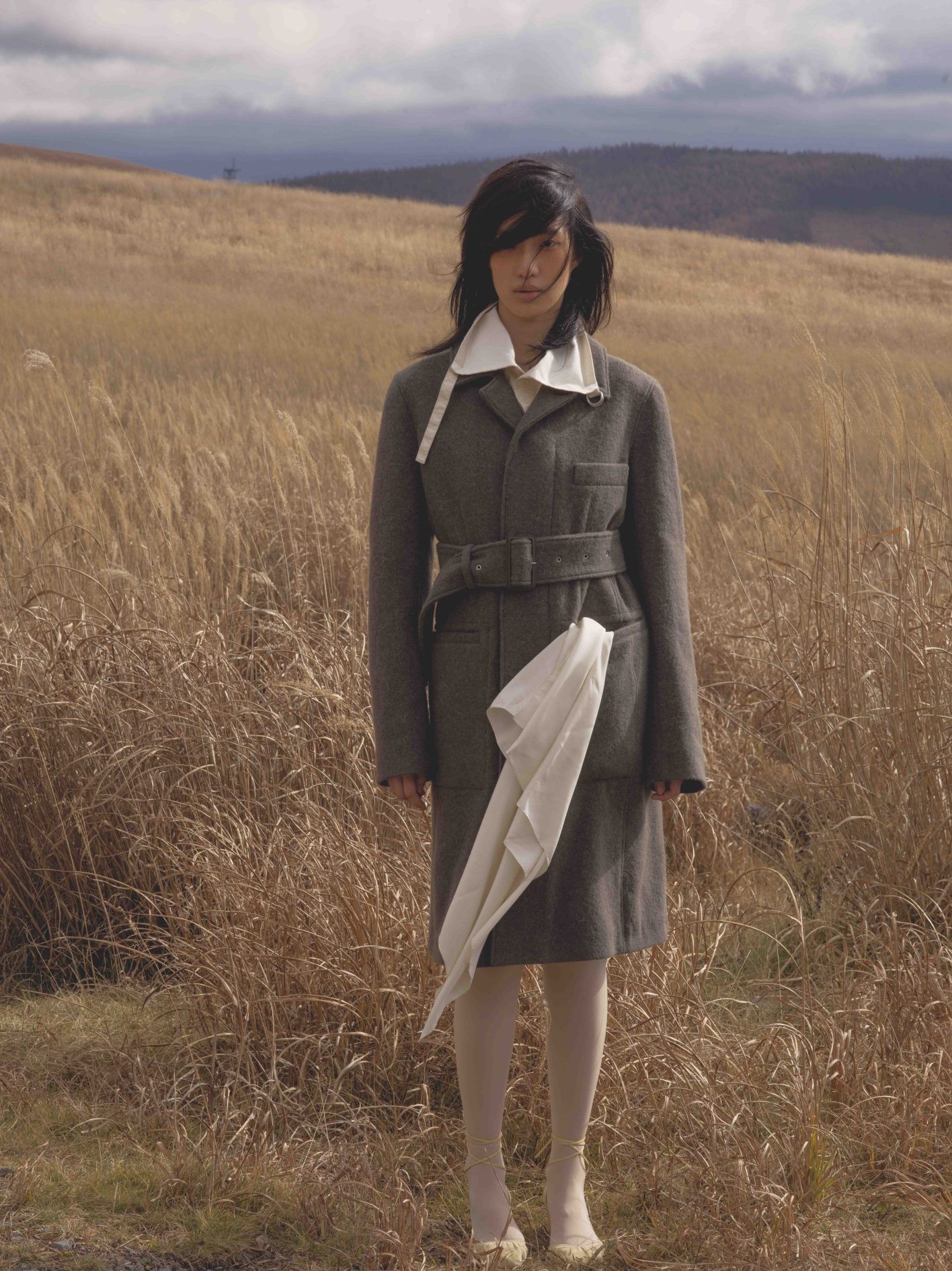 「自然から抽出したような中間色がいつも魅力的な〈ルメール〉。深く落ち着いたカーキ色に、静かな冬の森を想いました」(井伊さん)。「『枯野見』と称して、わざわざ枯れた野原を鑑賞する江戸っ子の余裕とセンスに触発されて、頰や唇にも侘びた色の遊びをほんのりと」(UDAさん)。マニッシュなベルト付きコート。首元からシャツの大きな襟を覗かせて。コート¥280,000、シャツ ¥122,000、シューズ ヒール5.5cm ¥109,000*参考価格(以上ルメール | エドストローム オフィス)/スカーフ、タイツ*スタイリスト私物